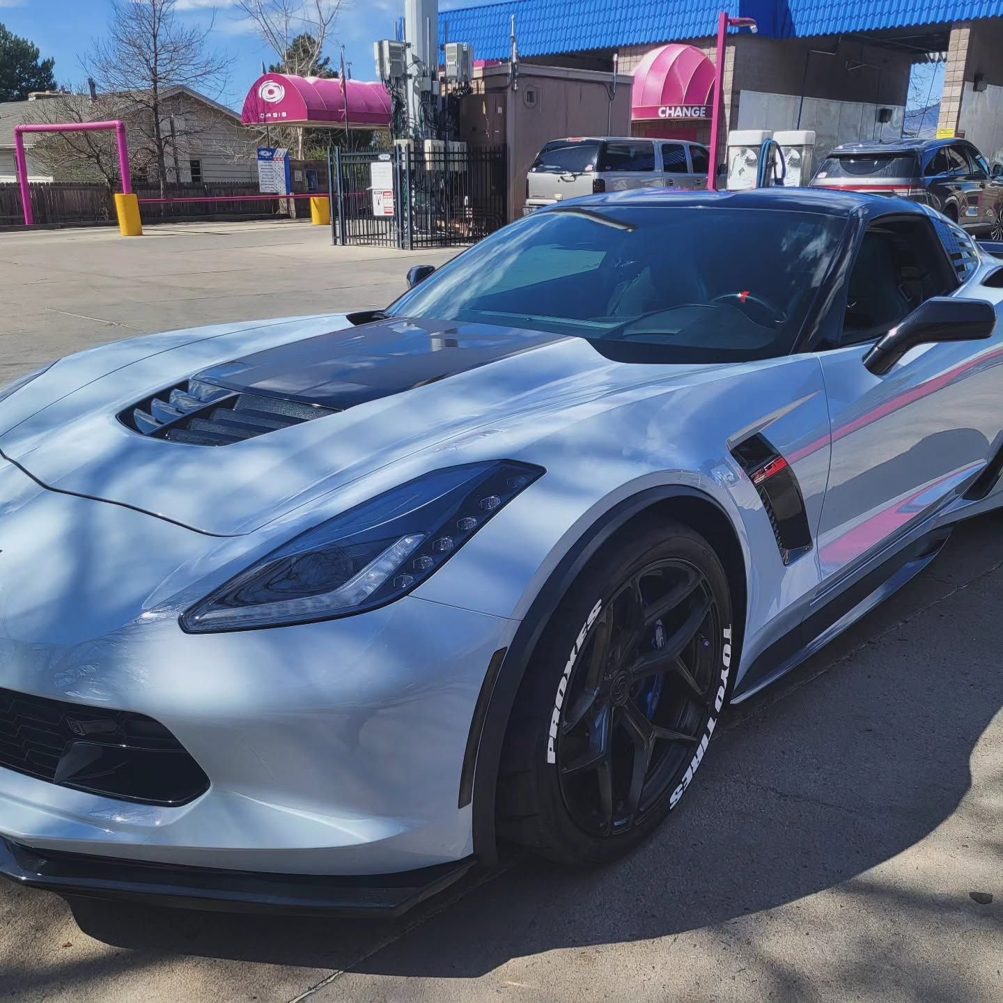 White sports car parked outdoors in bright sunlight, with a pink building and blue sky in the background