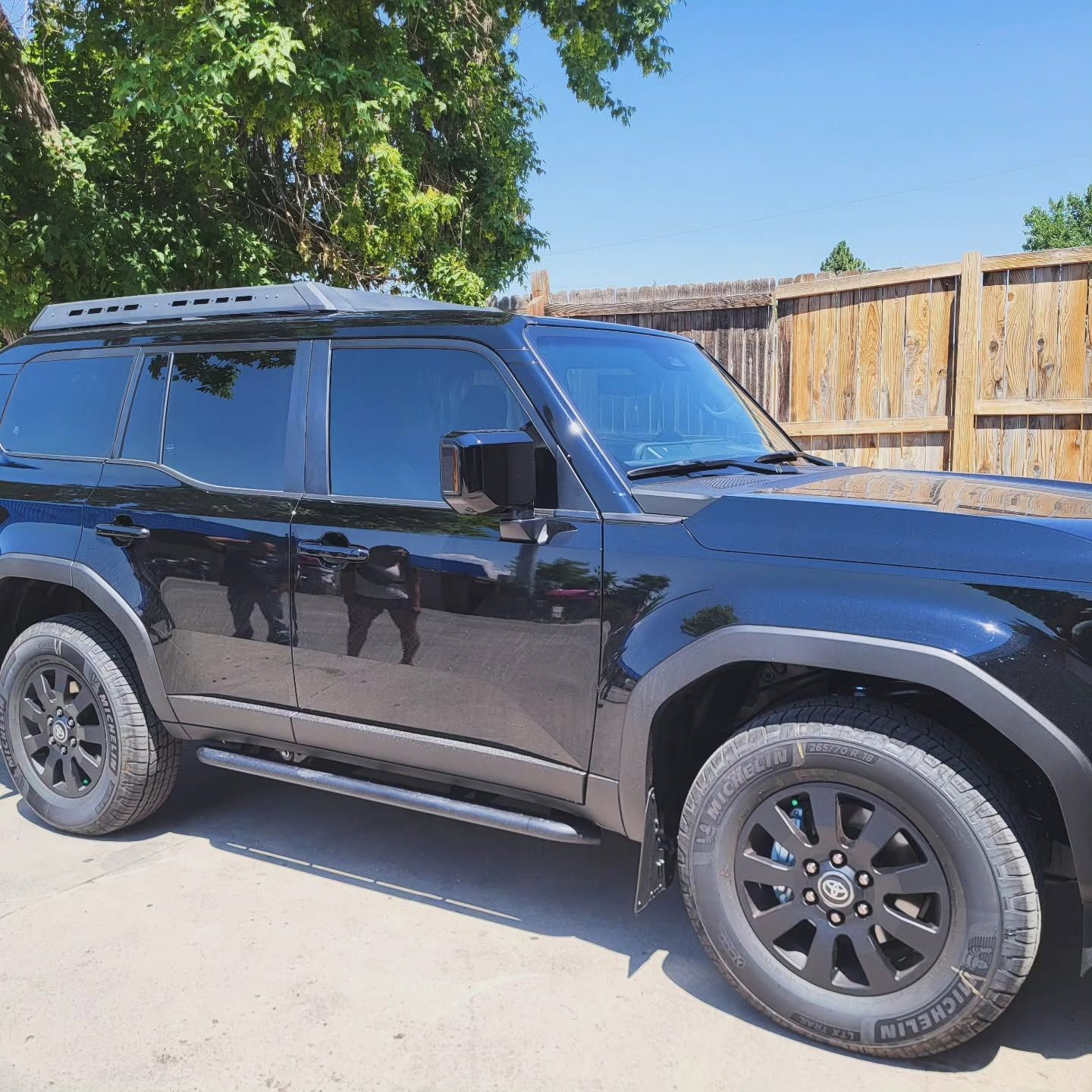 Black SUV parked on a sunny driveway beside a wooden fence and trees.