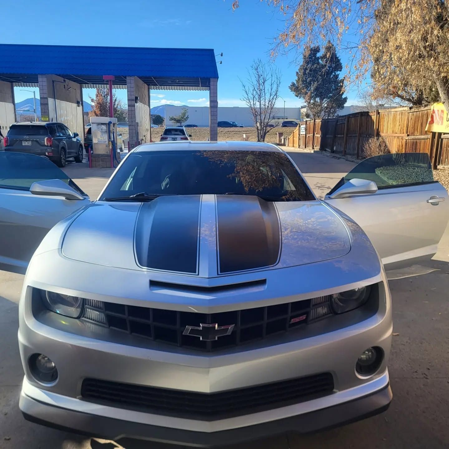 Front view of a silver Chevrolet Camaro with black racing stripes, parked outdoors with doors open.