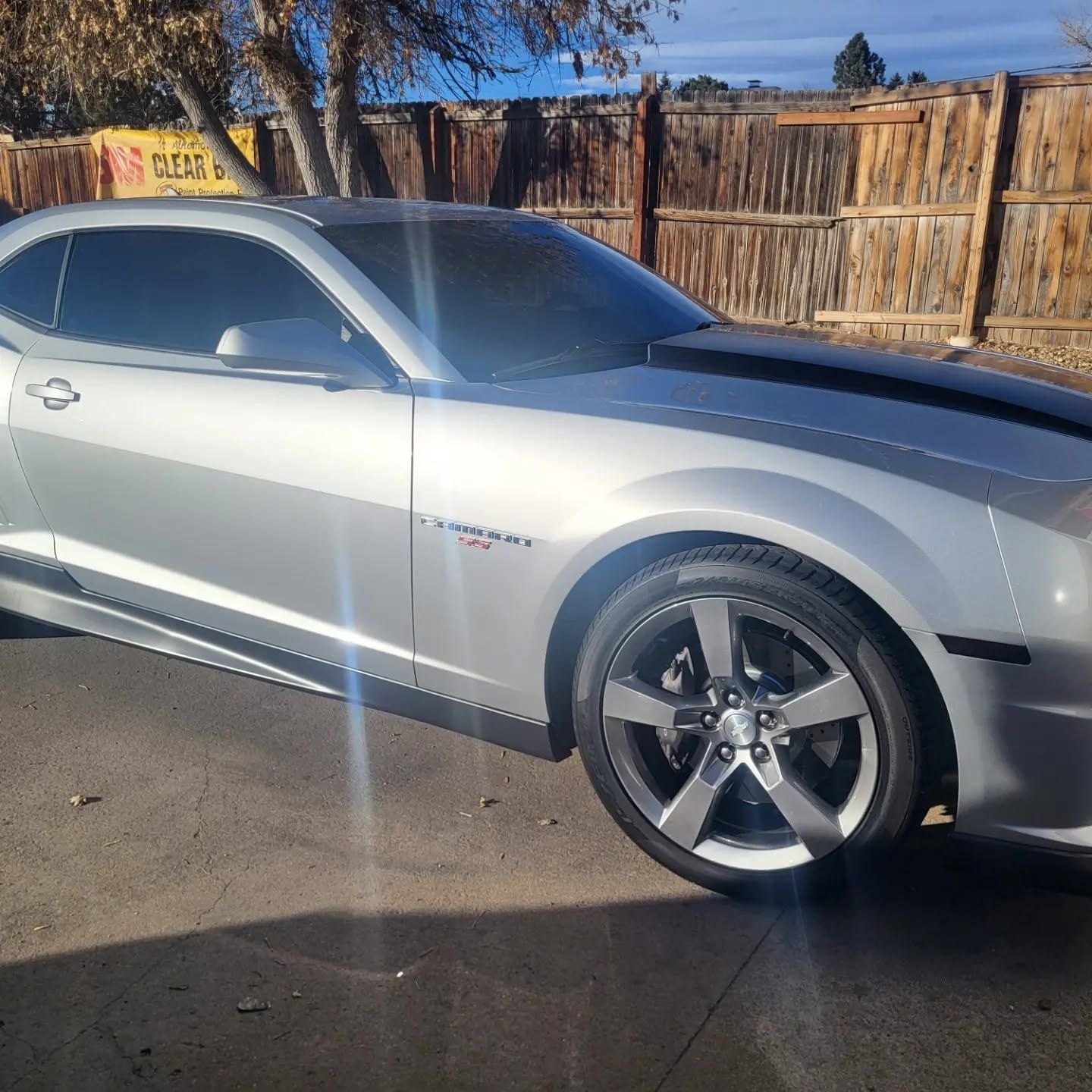 Silver sports coupe parked outdoors beside a wooden fence in bright sunlight.