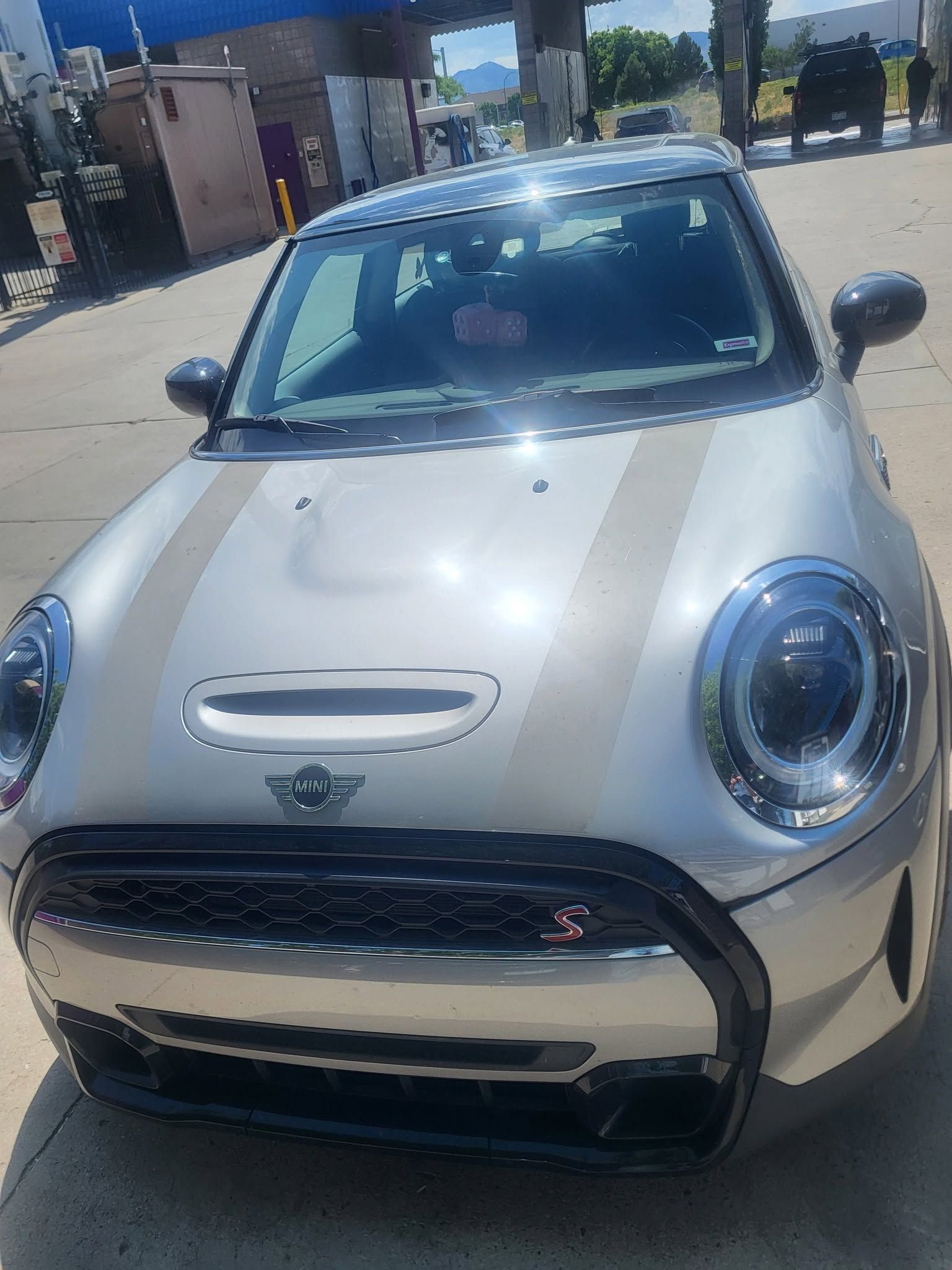 Silver Mini Cooper parked outdoors near a gas station, front view in bright sunlight