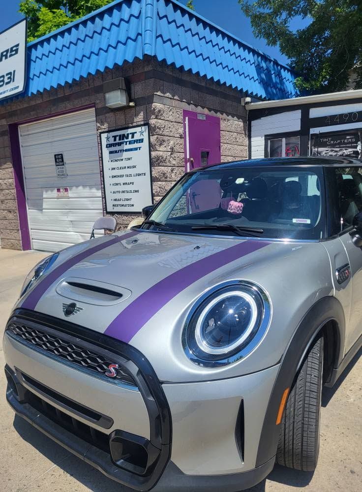 Silver Mini Cooper with purple racing stripes parked outside a small shop with a blue awning.