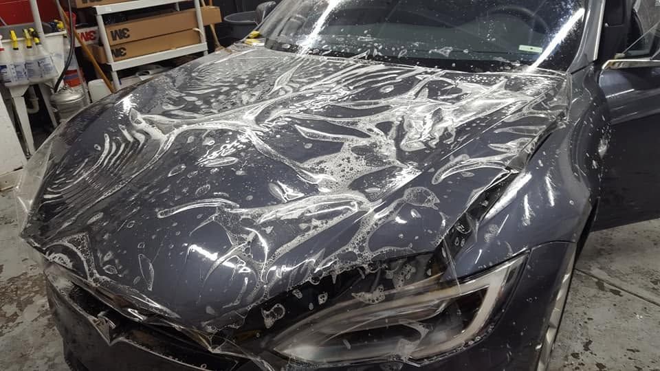 Damaged black car with shattered windshield and crumpled hood in a garage, covered in clear plastic