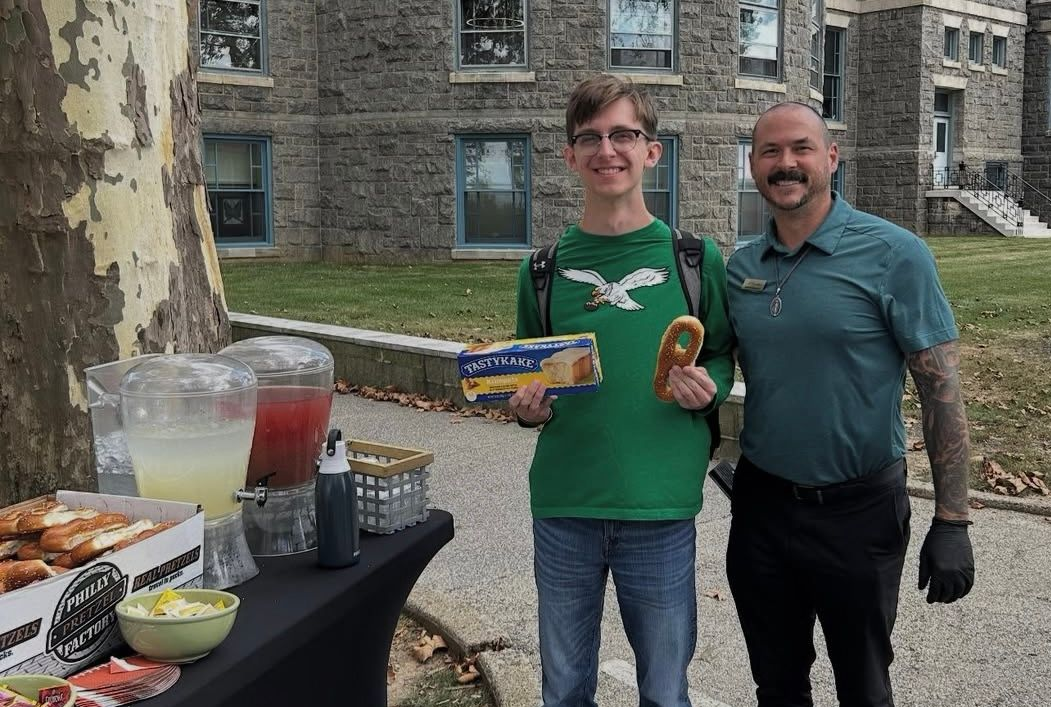 Two men posing with food and drink at a table outdoors in front of a stone building.