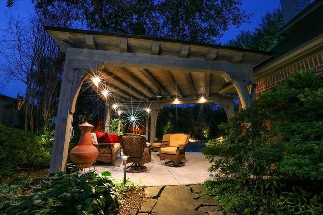 Outdoor seating area under a pergola at dusk, lit with lights, patio furniture, and lush greenery.