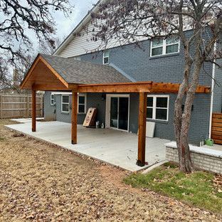 A gray brick house with a wooden patio cover over a concrete patio; a tree is on the right.
