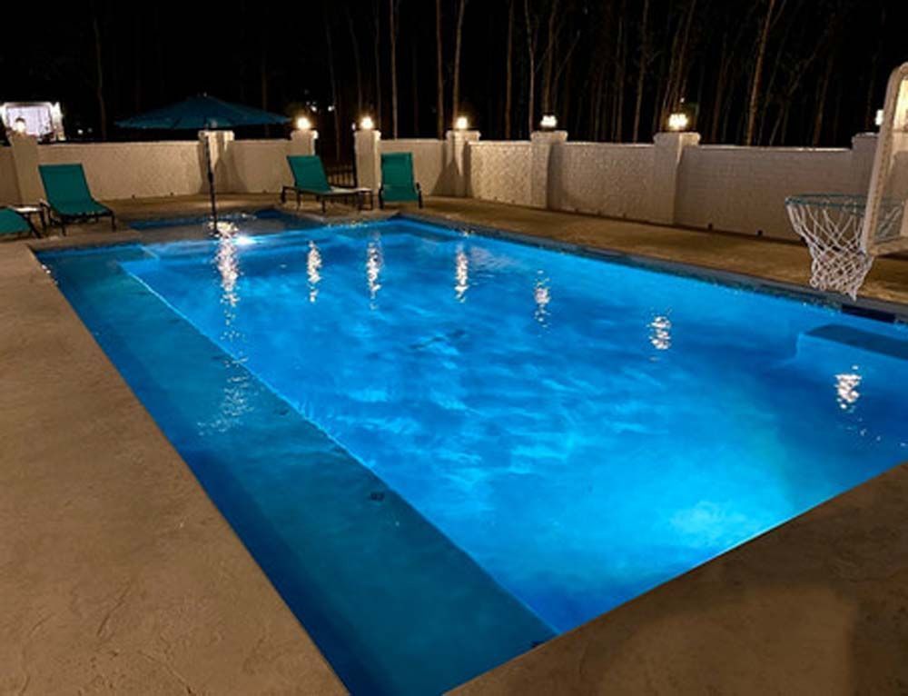 A nighttime view of a lit rectangular pool with blue water, surrounded by a concrete patio and white wall.