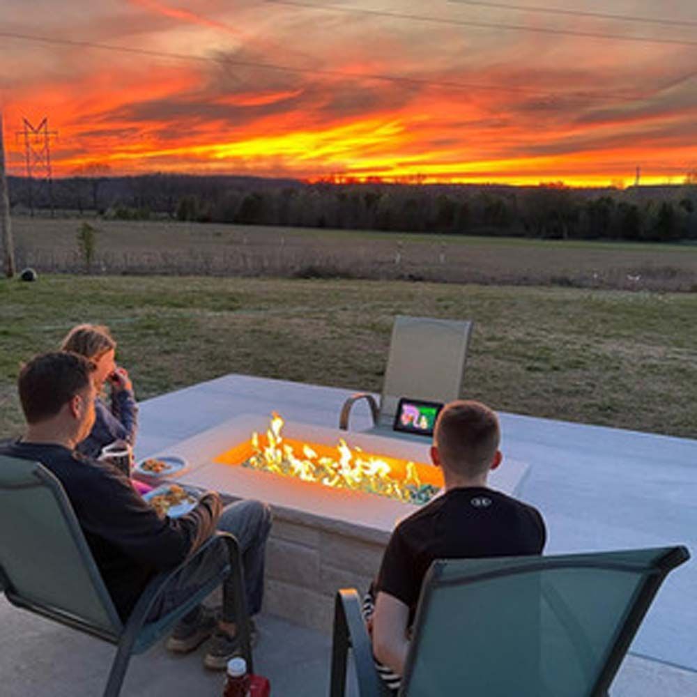 People around a fire pit at sunset. Orange sky, green chairs, field in background.