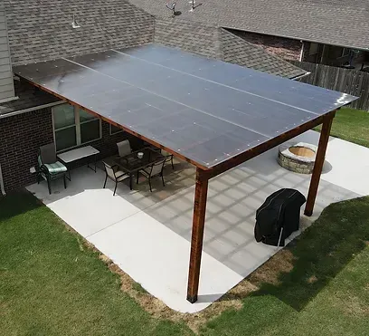Patio with wooden frame, clear roofing. Table, chairs, fire pit, and grill on a concrete slab.