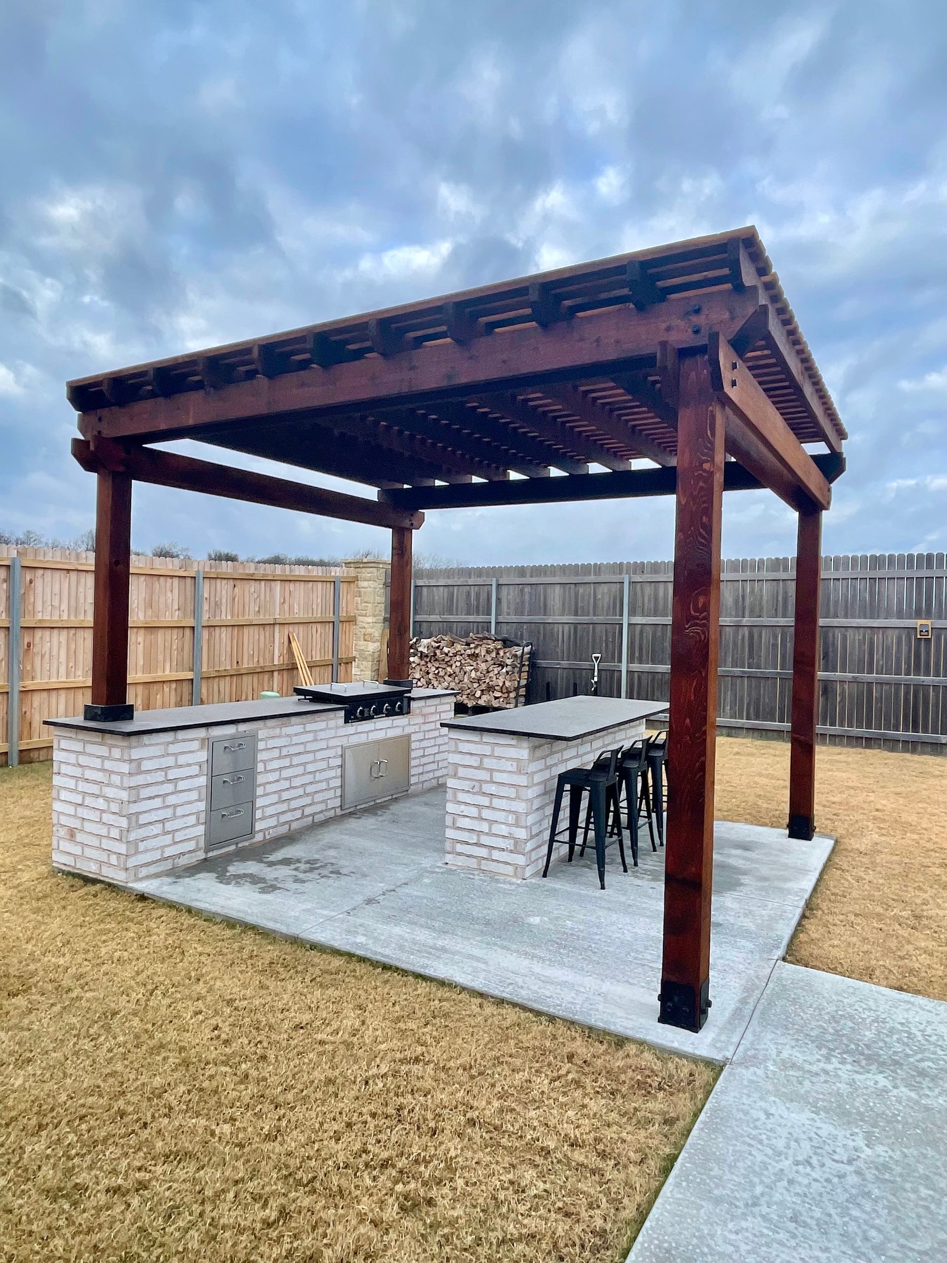 Outdoor kitchen under a brown pergola on a concrete patio. Features a grill, counter seating, and a wood pile.