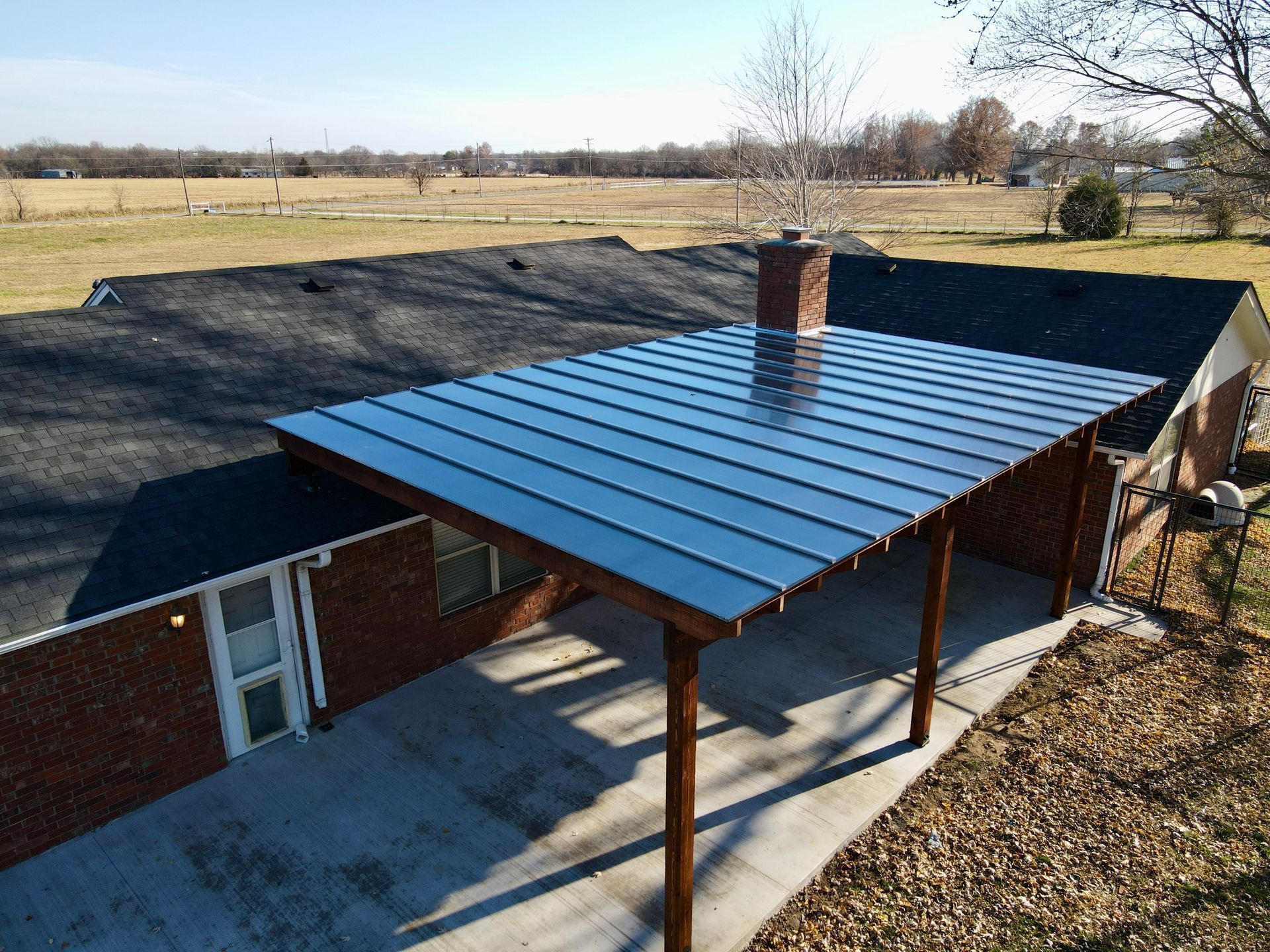 Carport with gray metal roof and wooden supports attached to a brick house on a concrete driveway.