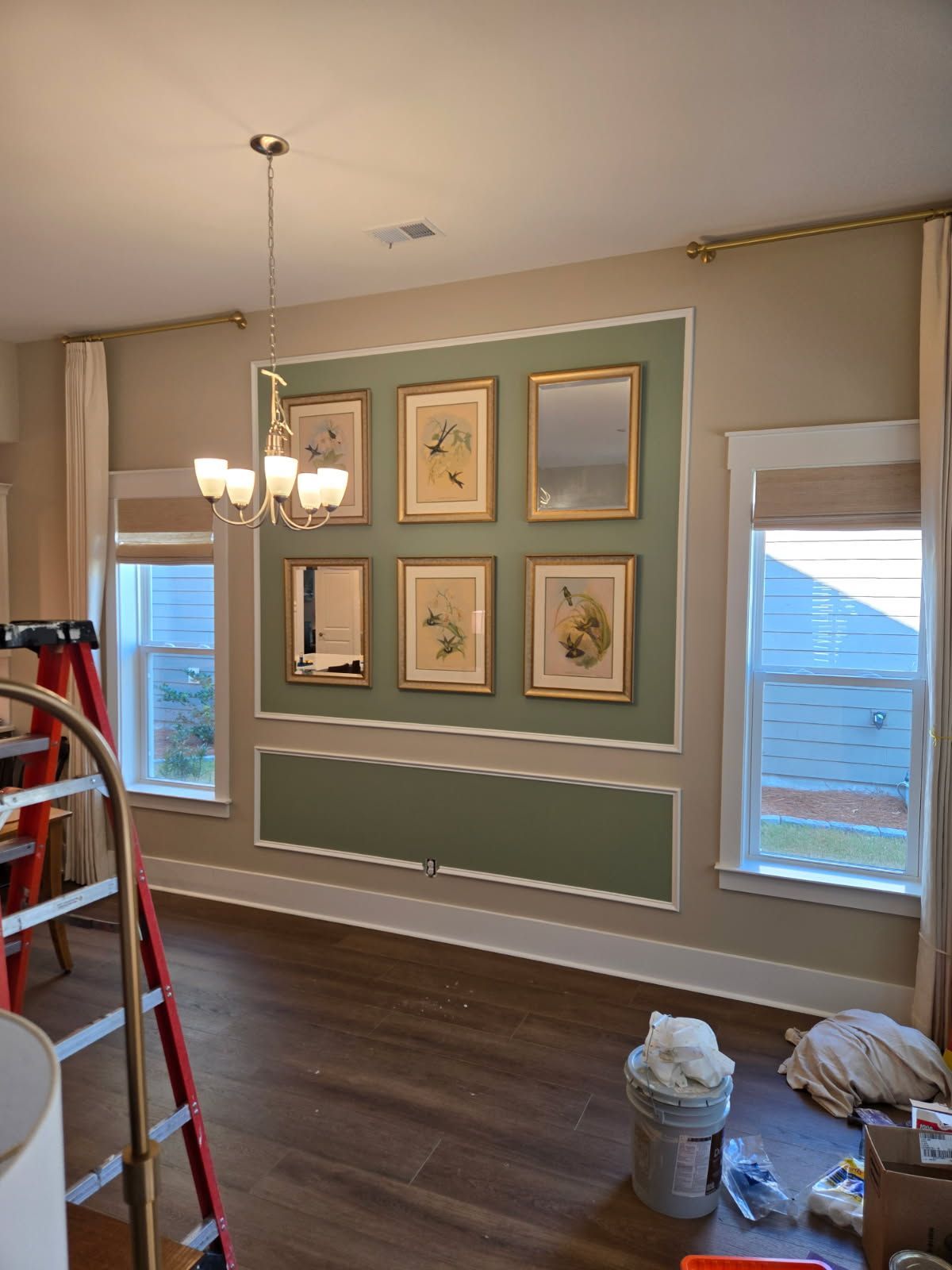 Dining room with sage green accent wall and framed art. Chandelier and windows visible.