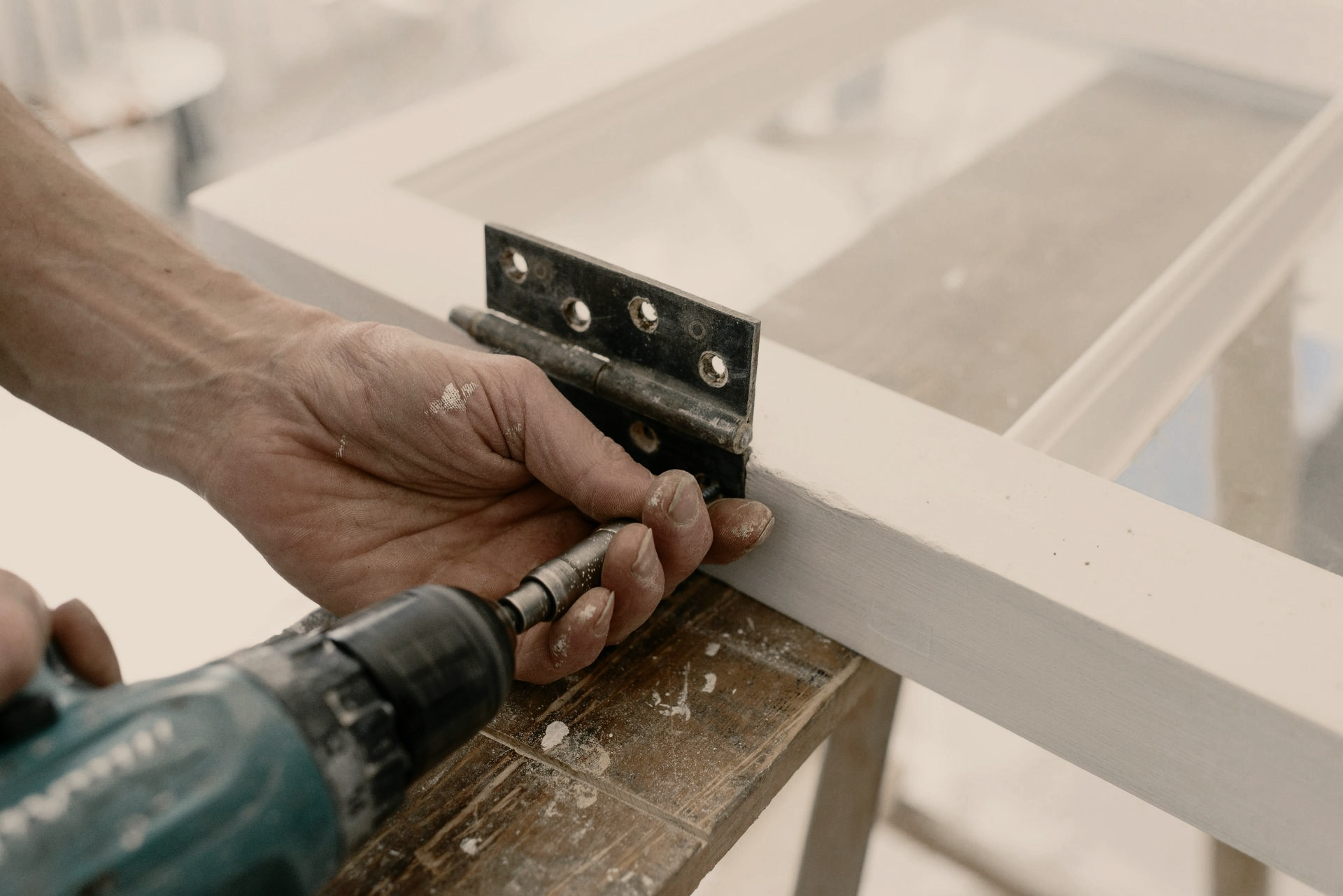 Person using a power drill to attach a hinge to a white-painted wooden frame.