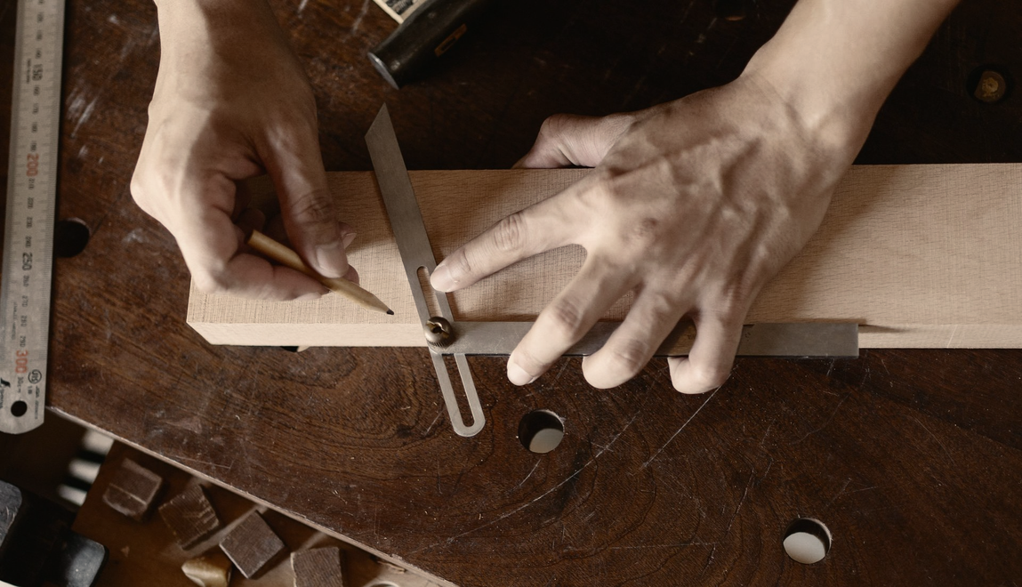 Hands using a square to measure and mark a line on a wooden plank on a workbench.