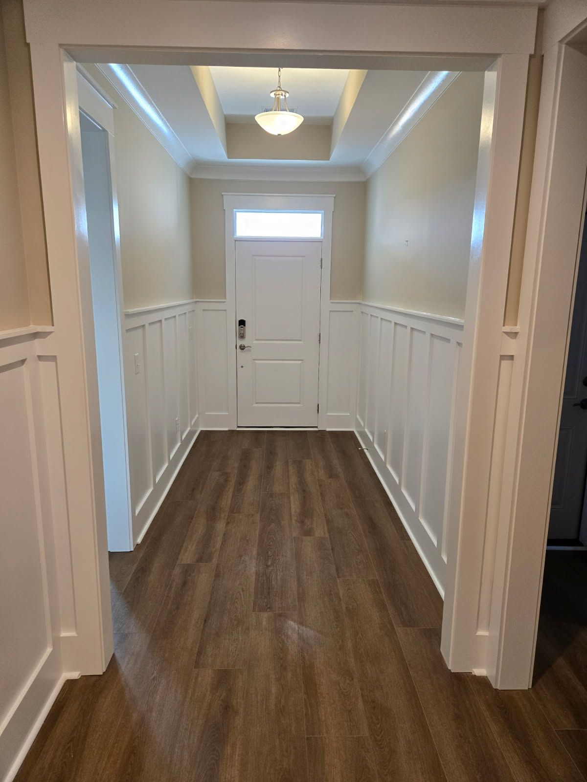 Hallway with white wainscoting, a white door, and dark wood-look flooring. A light fixture hangs from the ceiling.