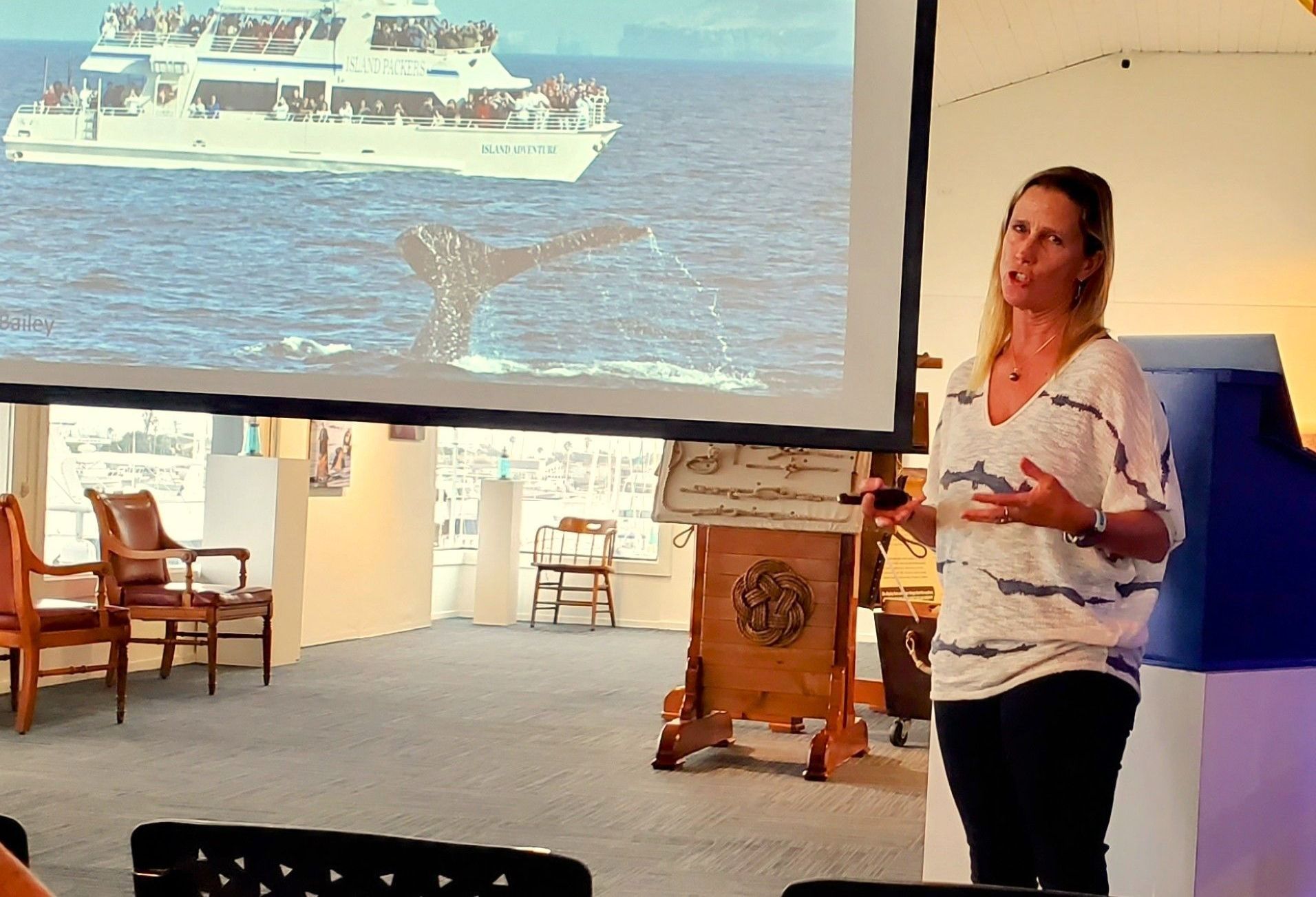 A presenter stands before a large screen displaying a boat at sea, gesturing while speaking to an audience.
