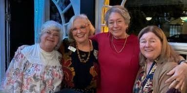 Four smiling people stand close together with arms around each other, posing for a photo in an indoor setting.