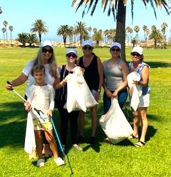 A group stands on a grassy field under palm trees, holding trash bags and tools, participating in a community cleanup.