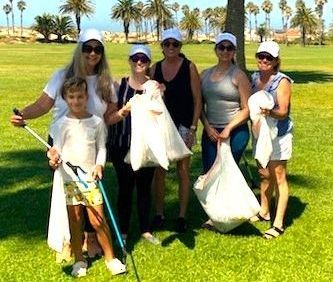 A group of people wearing white caps and holding white trash bags stand in a sunny, palm-lined park after a cleanup.
