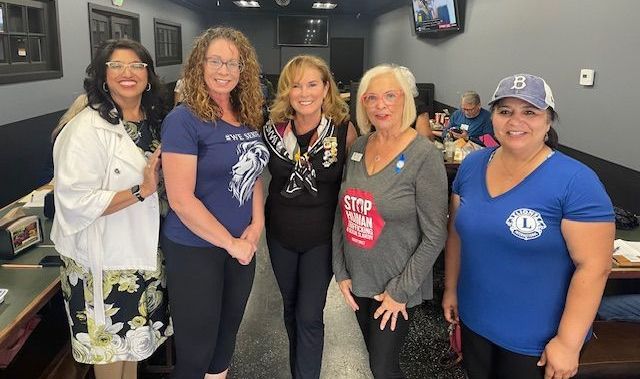 Five women stand smiling for a photo in a casual, dimly lit restaurant setting.