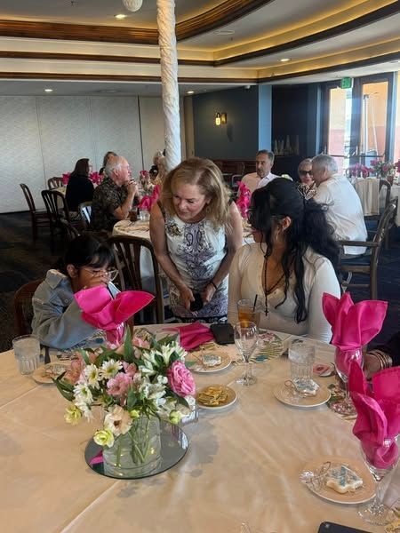 Girls seated at a banquet table while one leans in to speak, with pink napkins and flowers in a restaurant