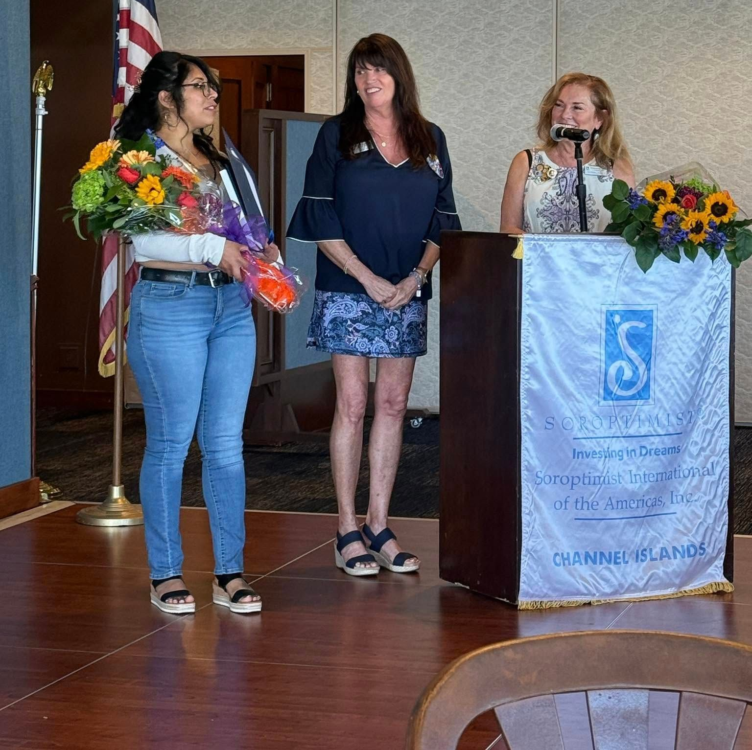 Three women stand on a stage beside a podium, with an American flag and flower arrangements.