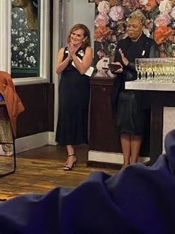 Two people in black clothing stand in a decorated indoor space, smiling and clapping, with glasses of champagne nearby.