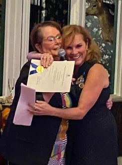 Two people embrace while one holds a certificate with a blue ribbon at an indoor event.