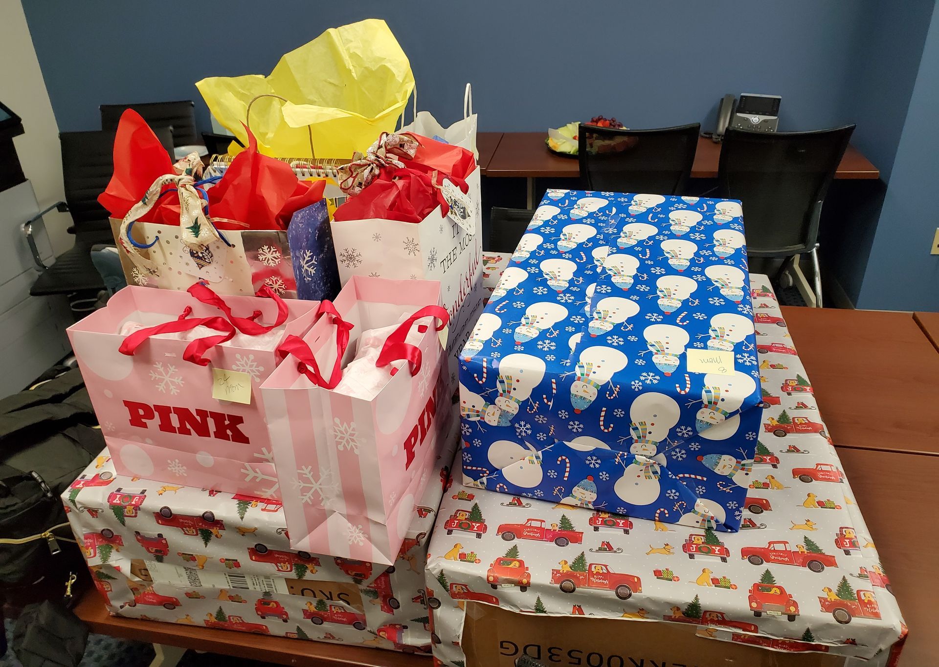 A collection of gifts, including PINK shopping bags and wrapped boxes with snowman and truck patterns, on a table.