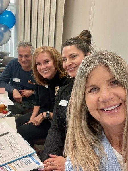 Four people smile at the camera during a gathering with blue balloons and papers on a table in the foreground.