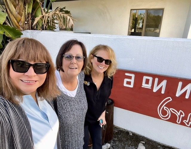 Three people wearing sunglasses stand side-by-side in front of a white wall with a partially visible red sign.