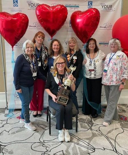 Seven people smile while posing in front of heart-shaped balloons at an event, with one person holding a small trophy.