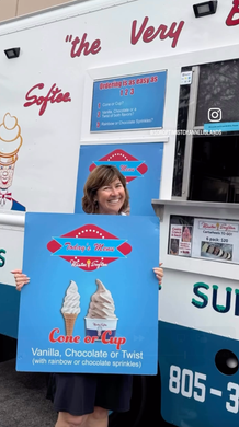 A person smiling while holding a large blue sign advertising ice cream in front of a soft-serve ice cream truck.