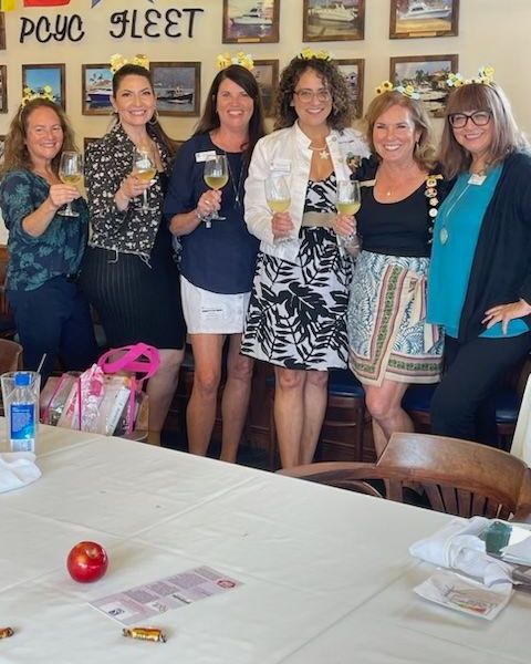 Six people in floral headbands stand in a row smiling and holding glasses of white wine in a room decorated with wall art.