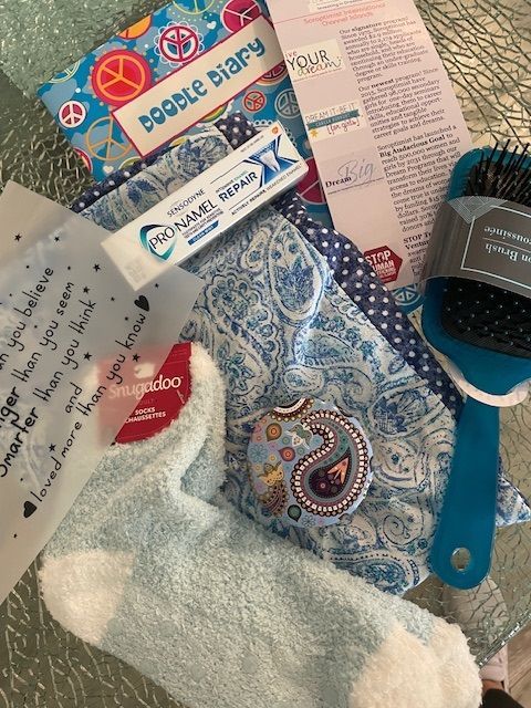 A collection of gift items including a blue doodle diary, ProNamel toothpaste, fuzzy socks, and a hairbrush on a table.