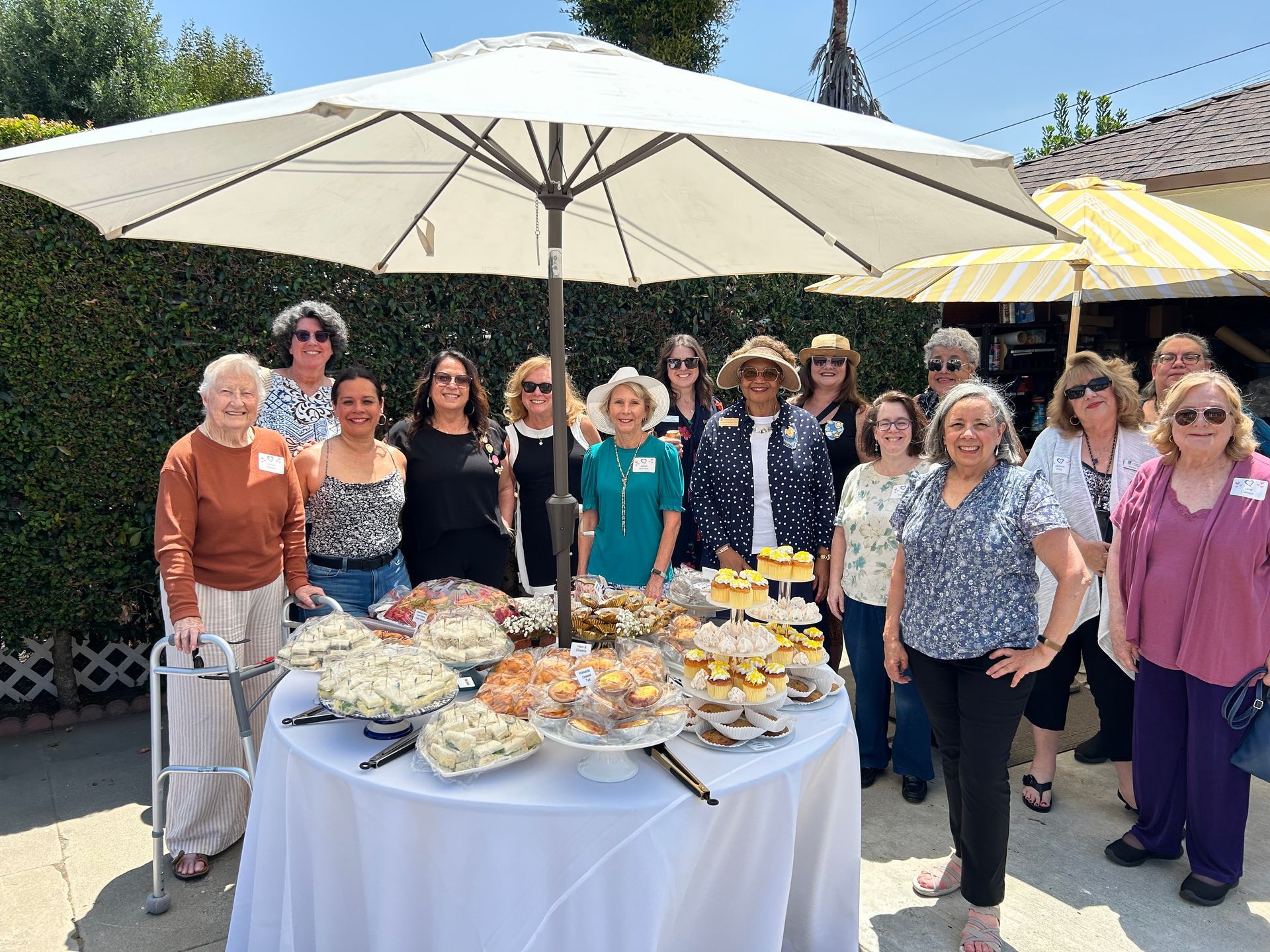 A diverse group of people gathers outdoors under umbrellas around a circular table laden with food at a sunny social event.