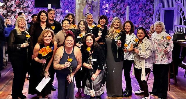 A group of people standing in a room with floral wallpaper, smiling and holding orange flowers for a celebratory event.
