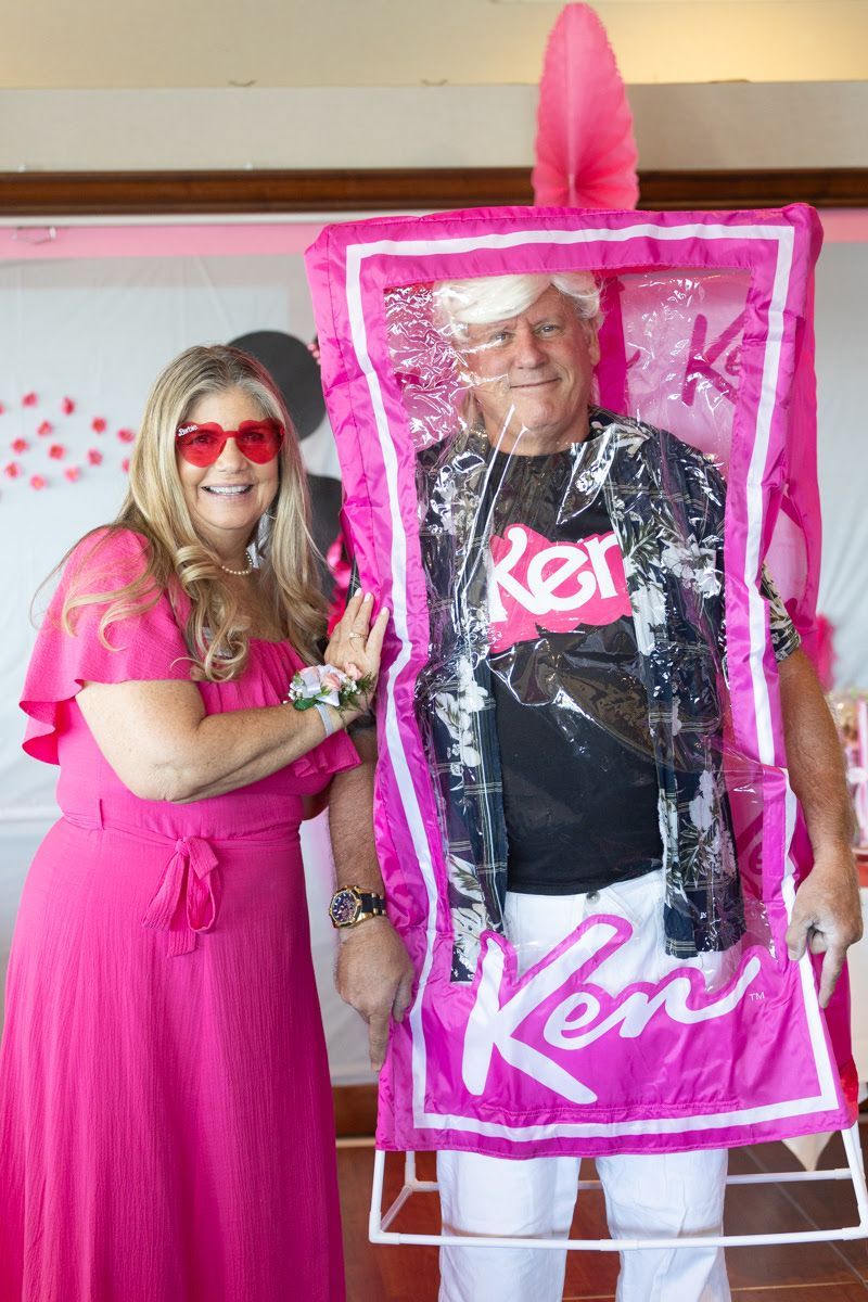 A person in a pink dress stands next to a person dressed as a Ken doll inside a pink doll box costume.
