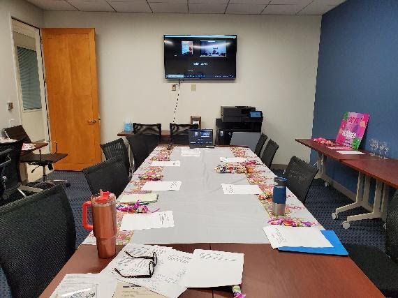 A conference table in an office, set with documents, a mug, and a wall-mounted display for a video meeting.