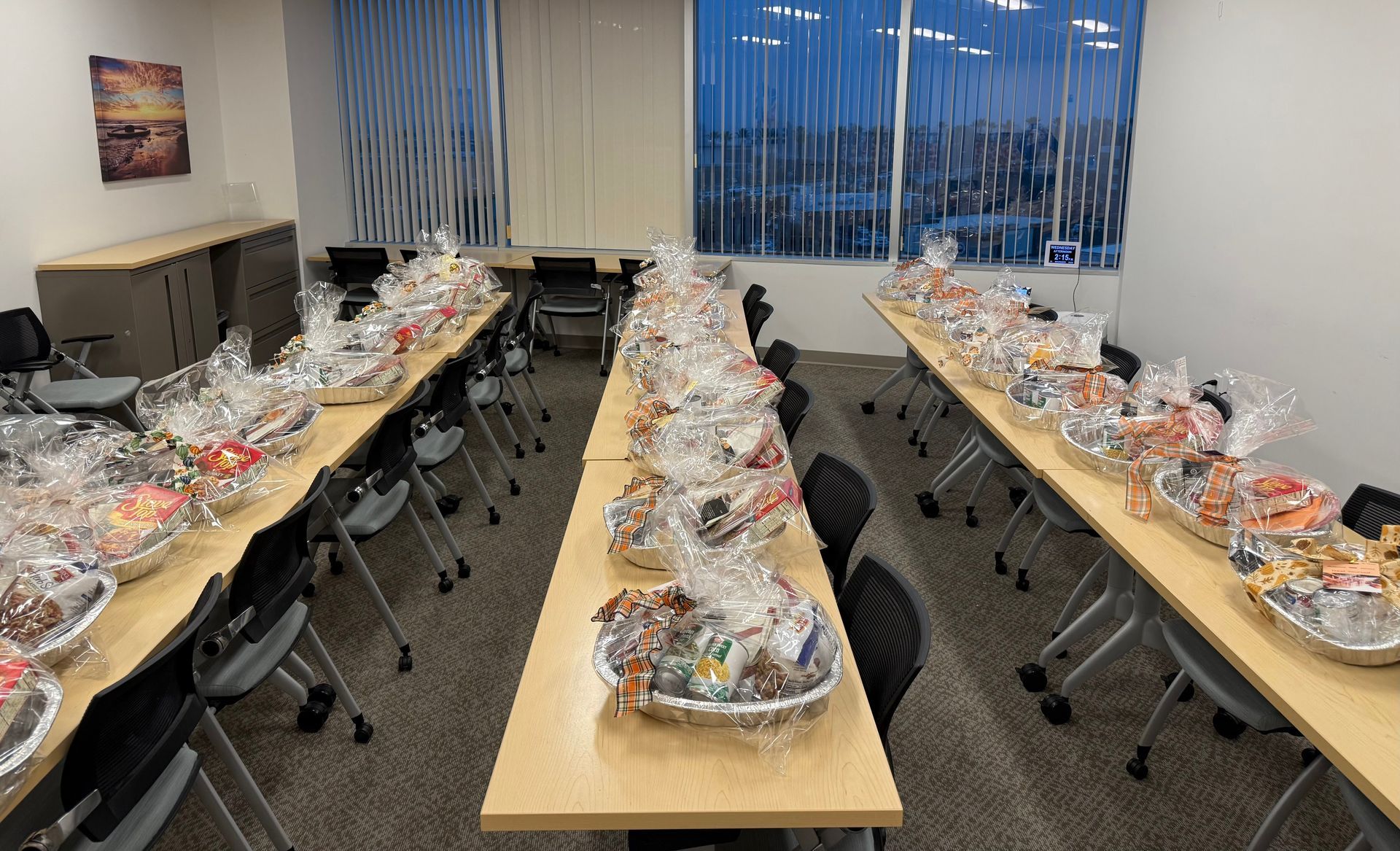 Rows of gift baskets wrapped in clear cellophane are arranged on tables in a brightly lit office meeting room.