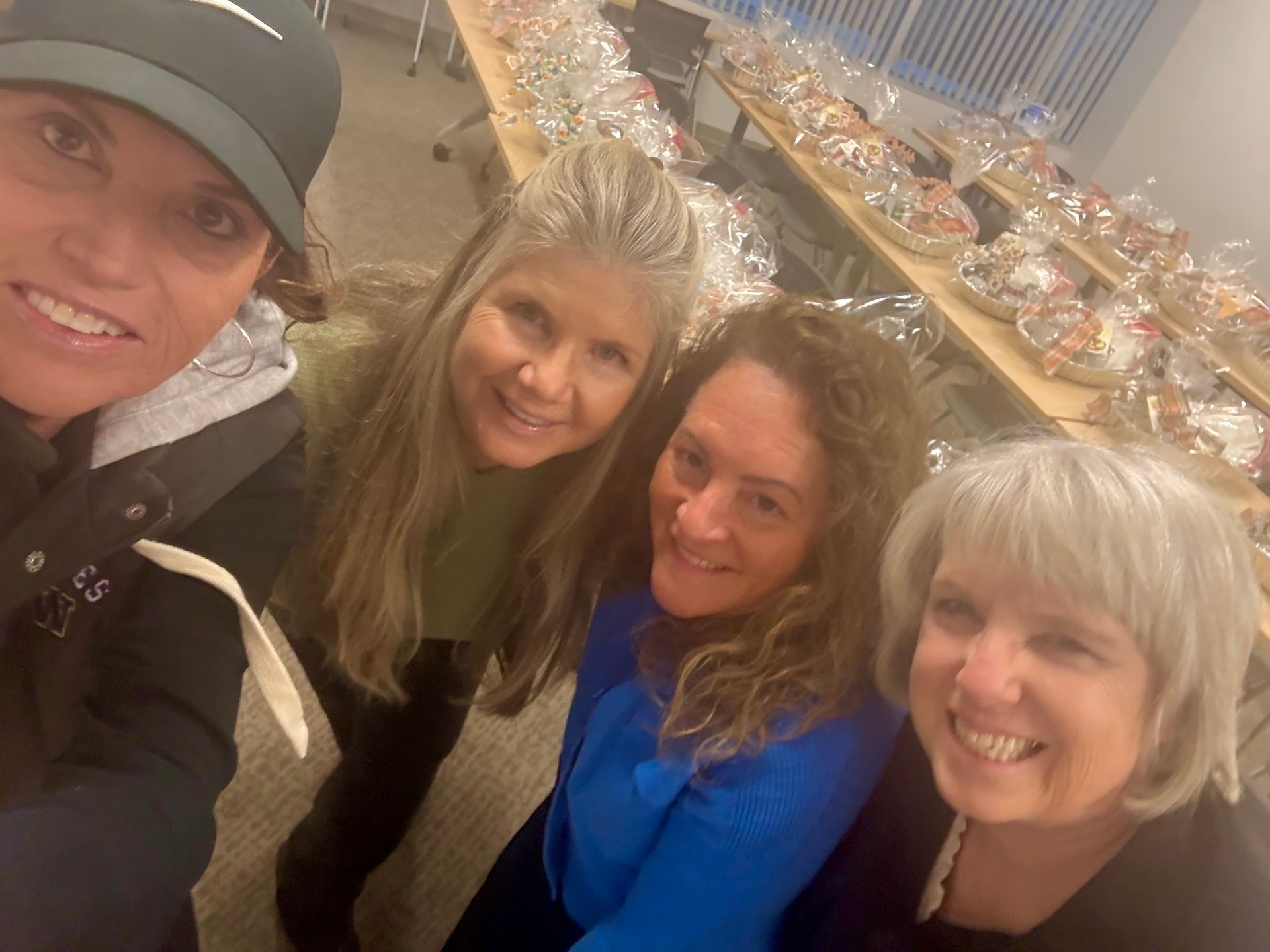Four people smile for a selfie in front of tables filled with wrapped gift baskets.