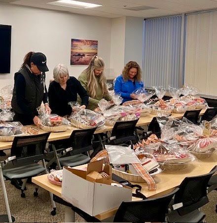 Four people working in an office setting, assembling and wrapping gift baskets on a long table.