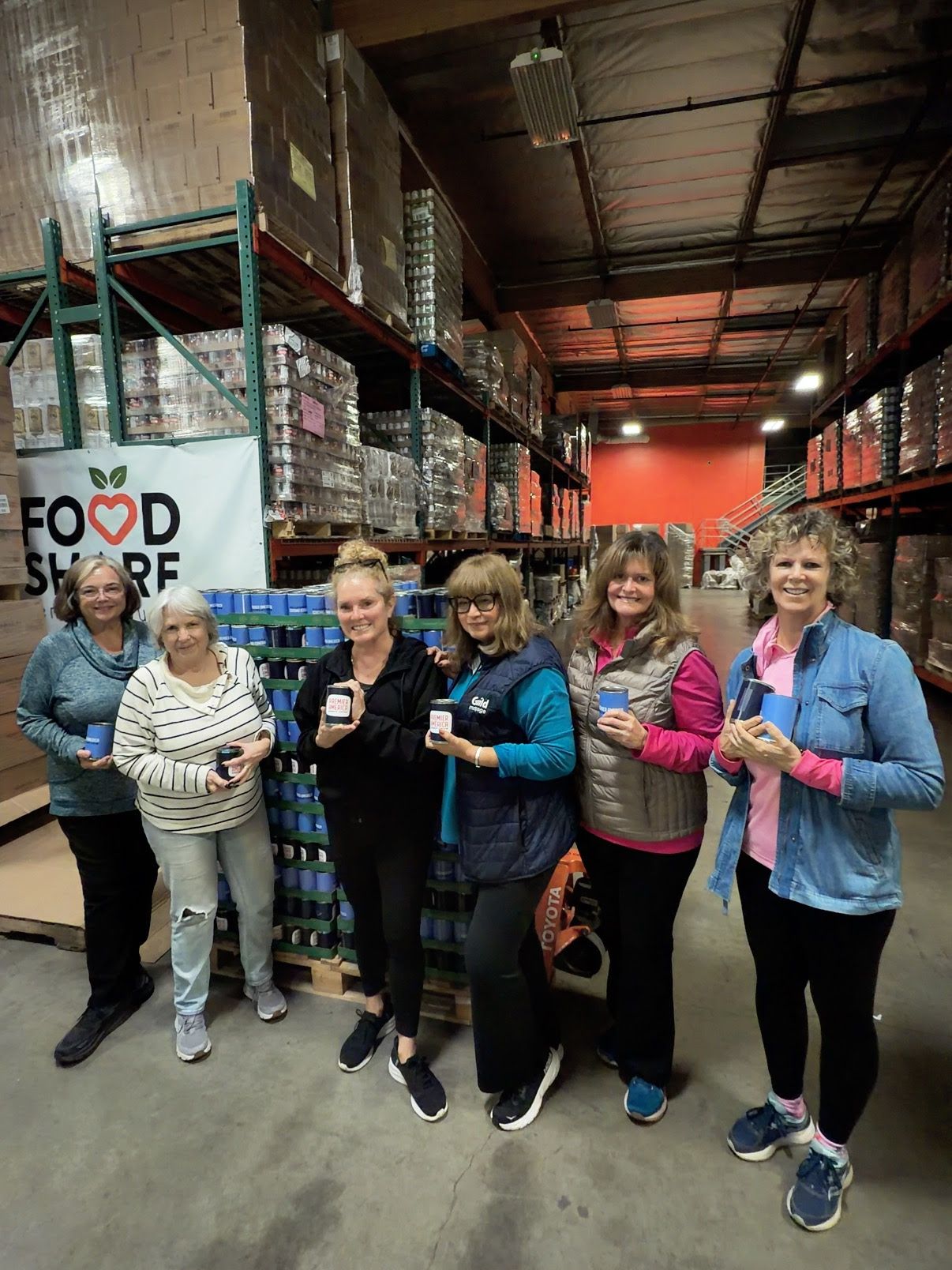Six volunteers stand in a warehouse in front of food supply shelves, holding small items and smiling for the camera.