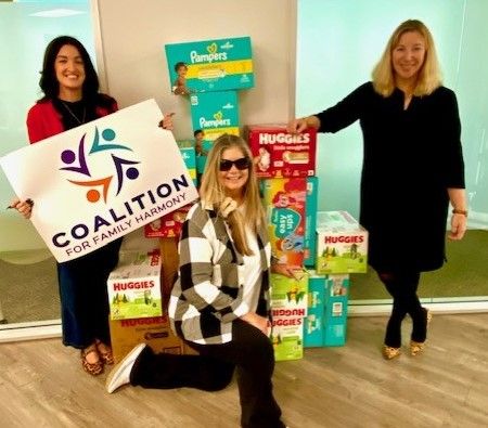 Three women pose with stacked diaper boxes and a Coalition for Community Care sign.