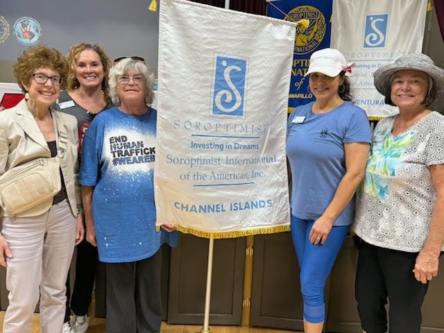 Five people stand holding a Soroptimist International of the Americas banner in a meeting room.