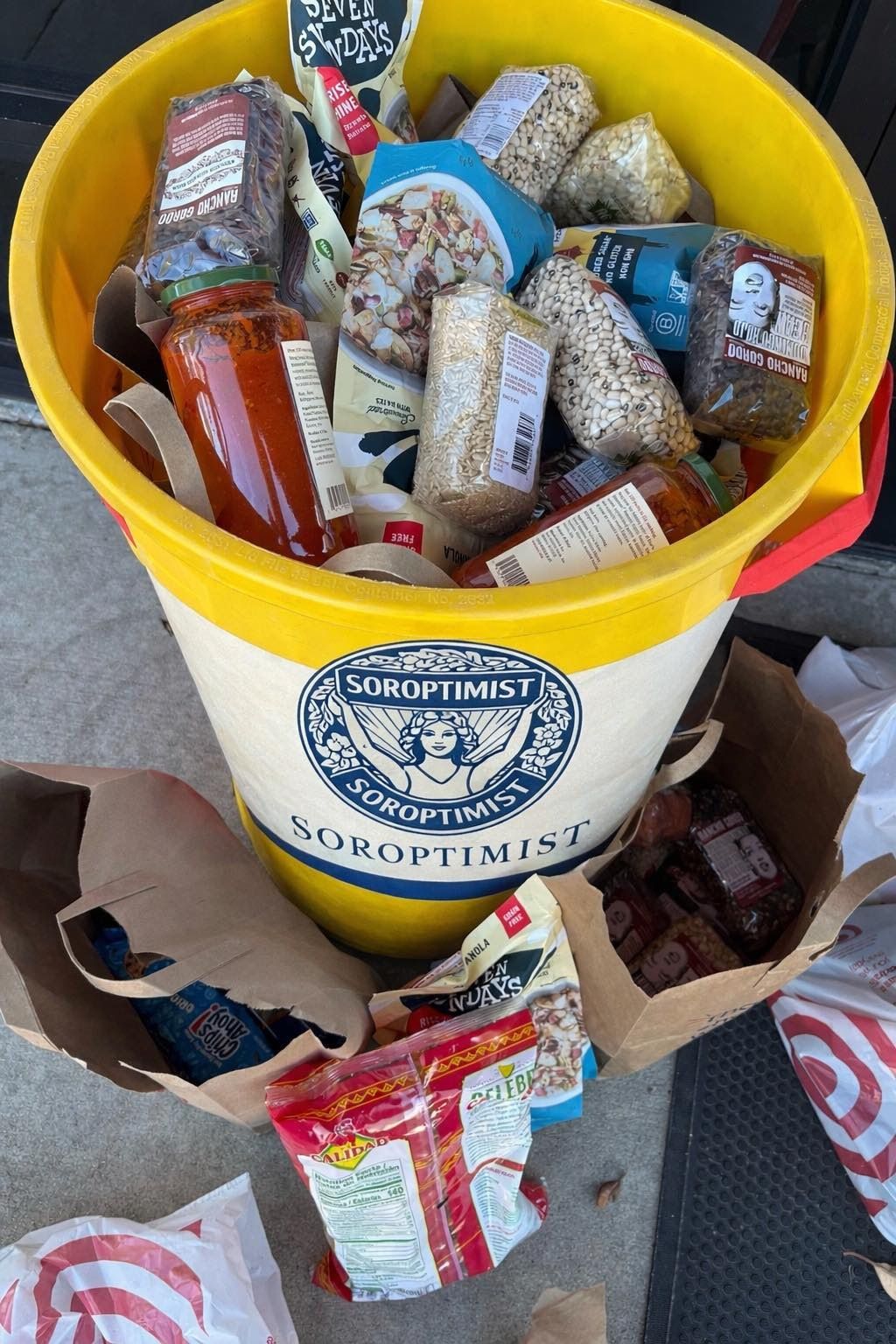 A yellow Soroptimist bucket and paper bags filled with assorted groceries and dry food items on a concrete surface.