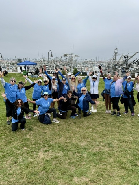 People in blue shirts pose with arms raised on a grassy field near the water. Women's Walk and Expo on Saturday, May 9, 2026.
