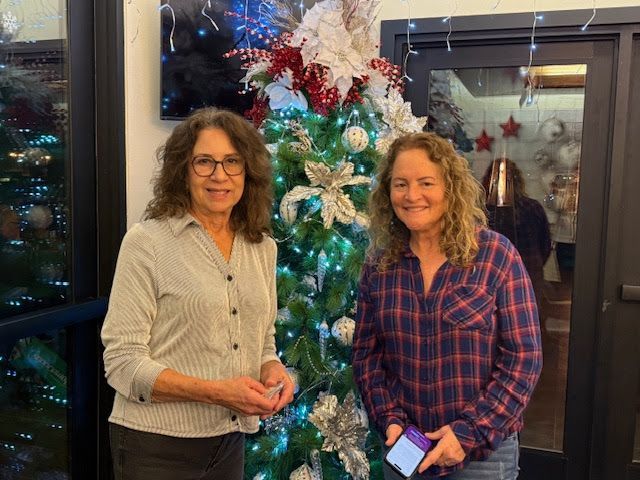 Two people smile while standing in front of a decorated Christmas tree in an indoor setting.