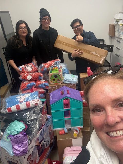 Four people smile while standing behind a collection of toys, gifts, and boxes in an office, likely for a donation drive.