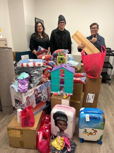 Three people stand behind a large collection of toys, gifts, and suitcases packed for donation in an indoor office space.